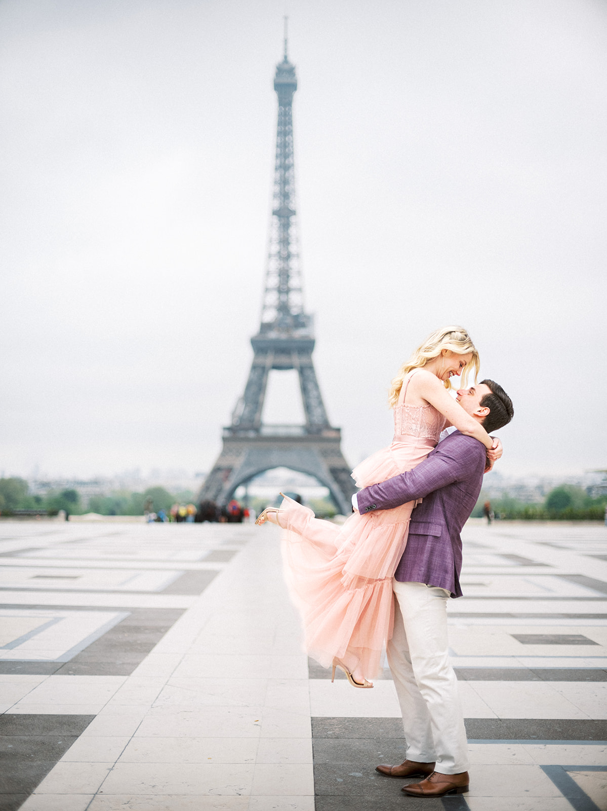 Man picks up woman in front of the Eiffel Tower as he looks at her.