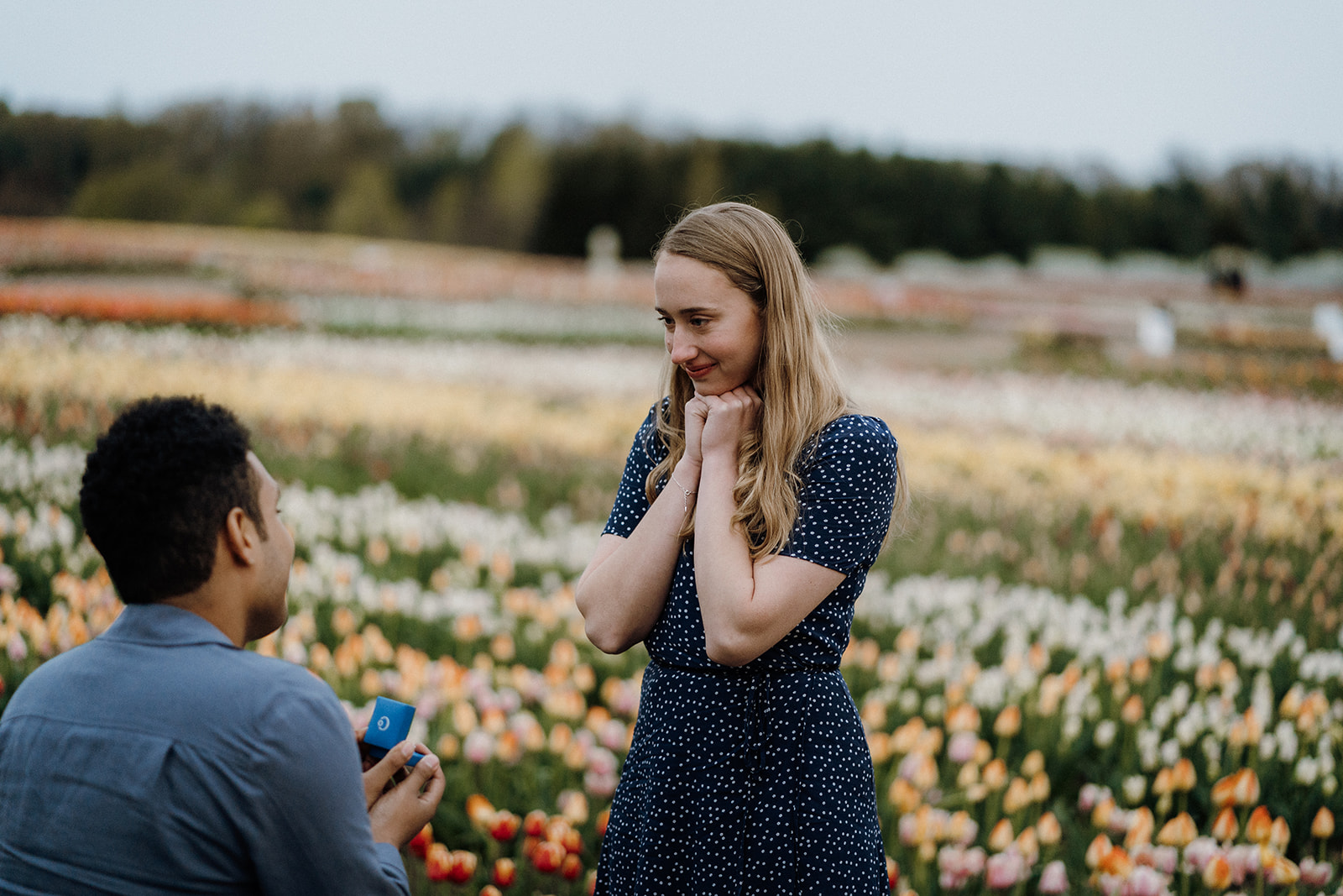 Man kneeling in tulips while his girlfriend holds her hands together underneath her chin.