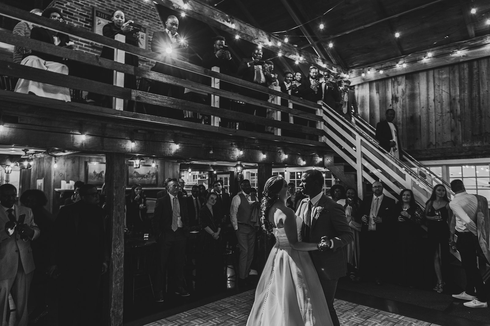 a black and white image of a bride and groom's first dance in an old barn with twinkle lights 