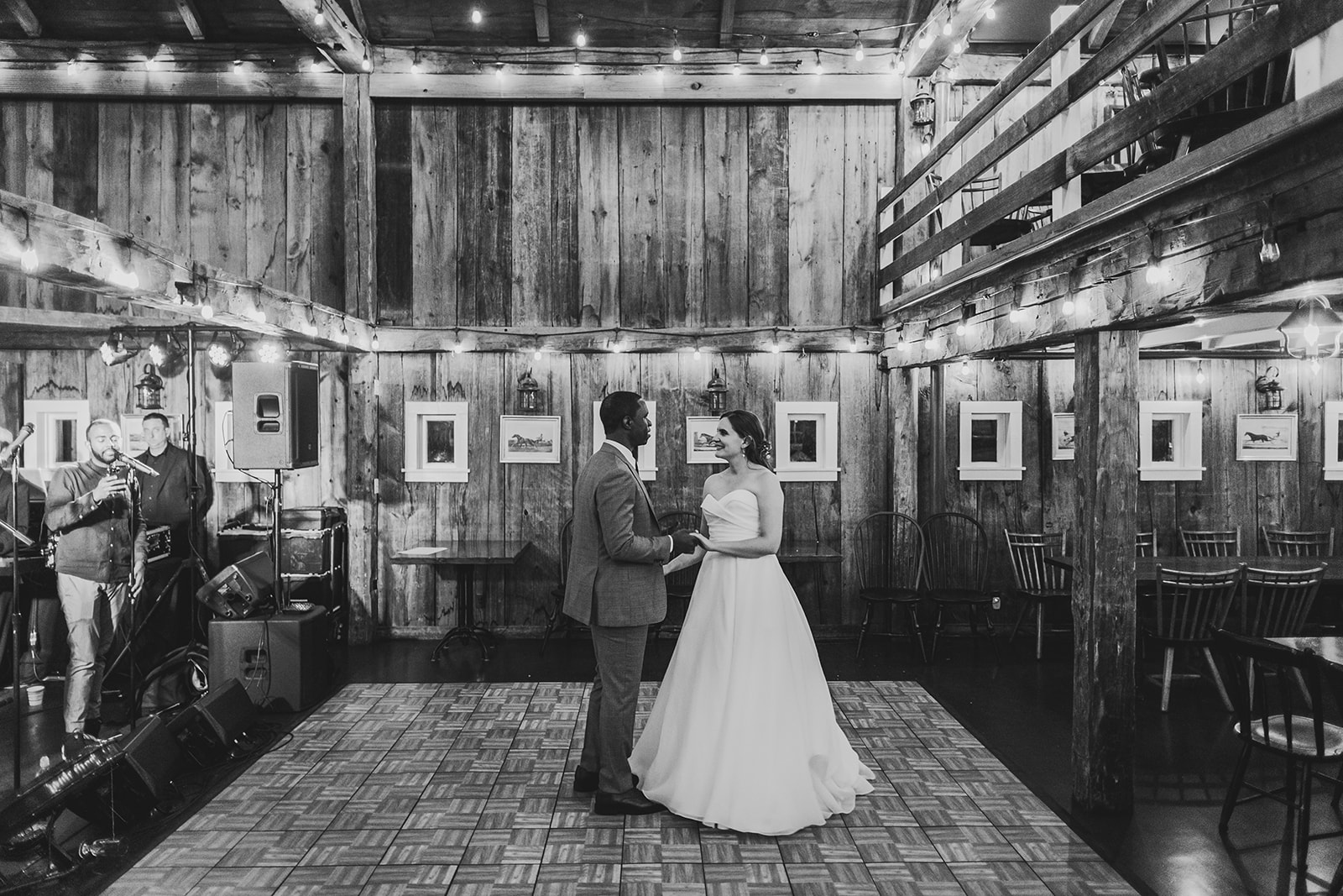 a black and white image of a bride and groom's first dance in an old barn with twinkle lights 