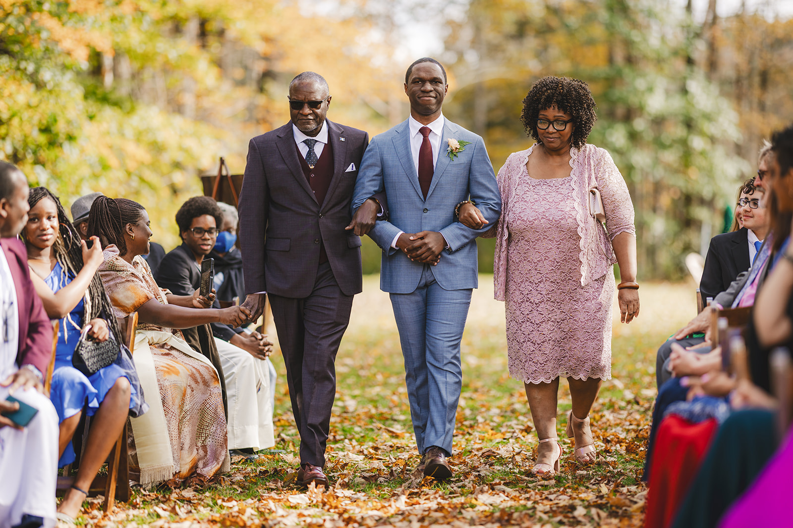 a groom in a blue suit is walked down the aisle by his parents at an outdoor wedding in the fall