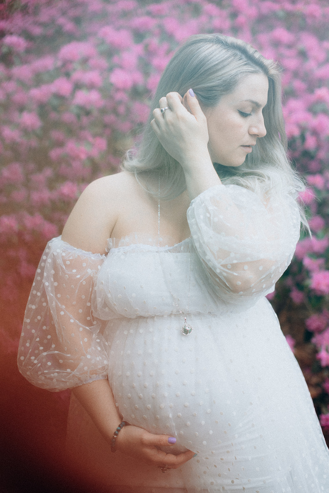 Pregnant Anais in front of pink flowers bush holding her baby bump and adjusting her hair