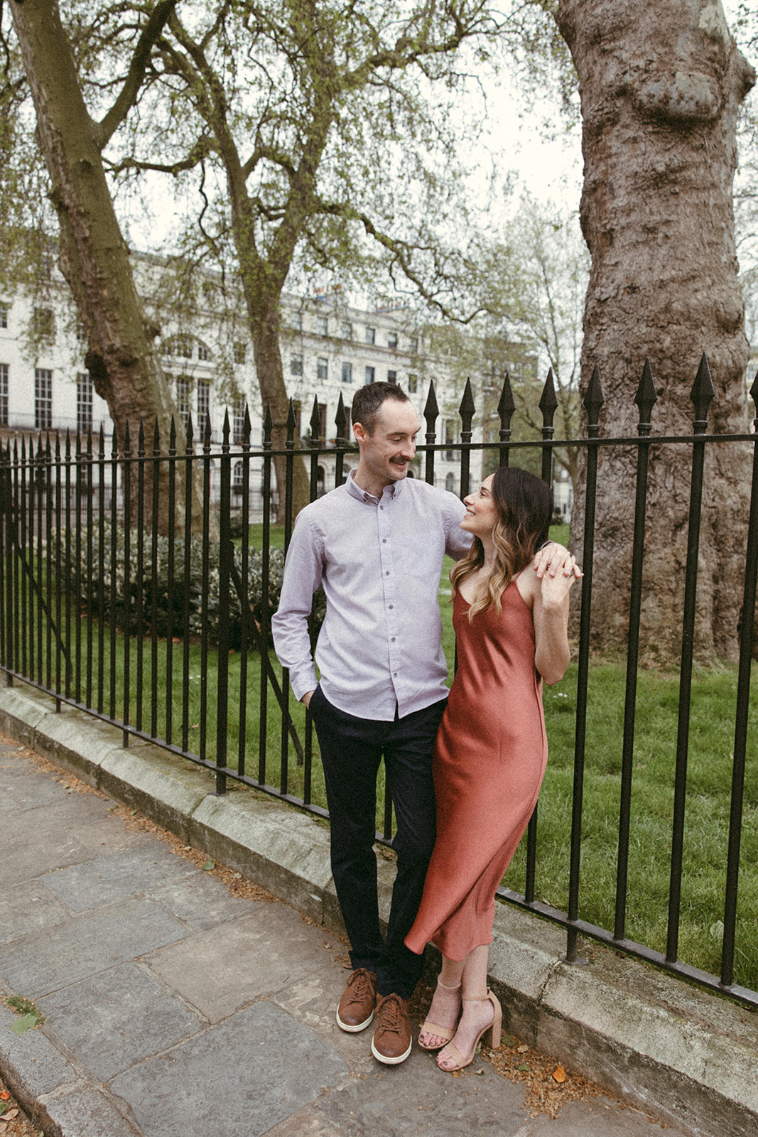 A London engagement session couple leans against a black wrought iron fence at Regent’s Park.
