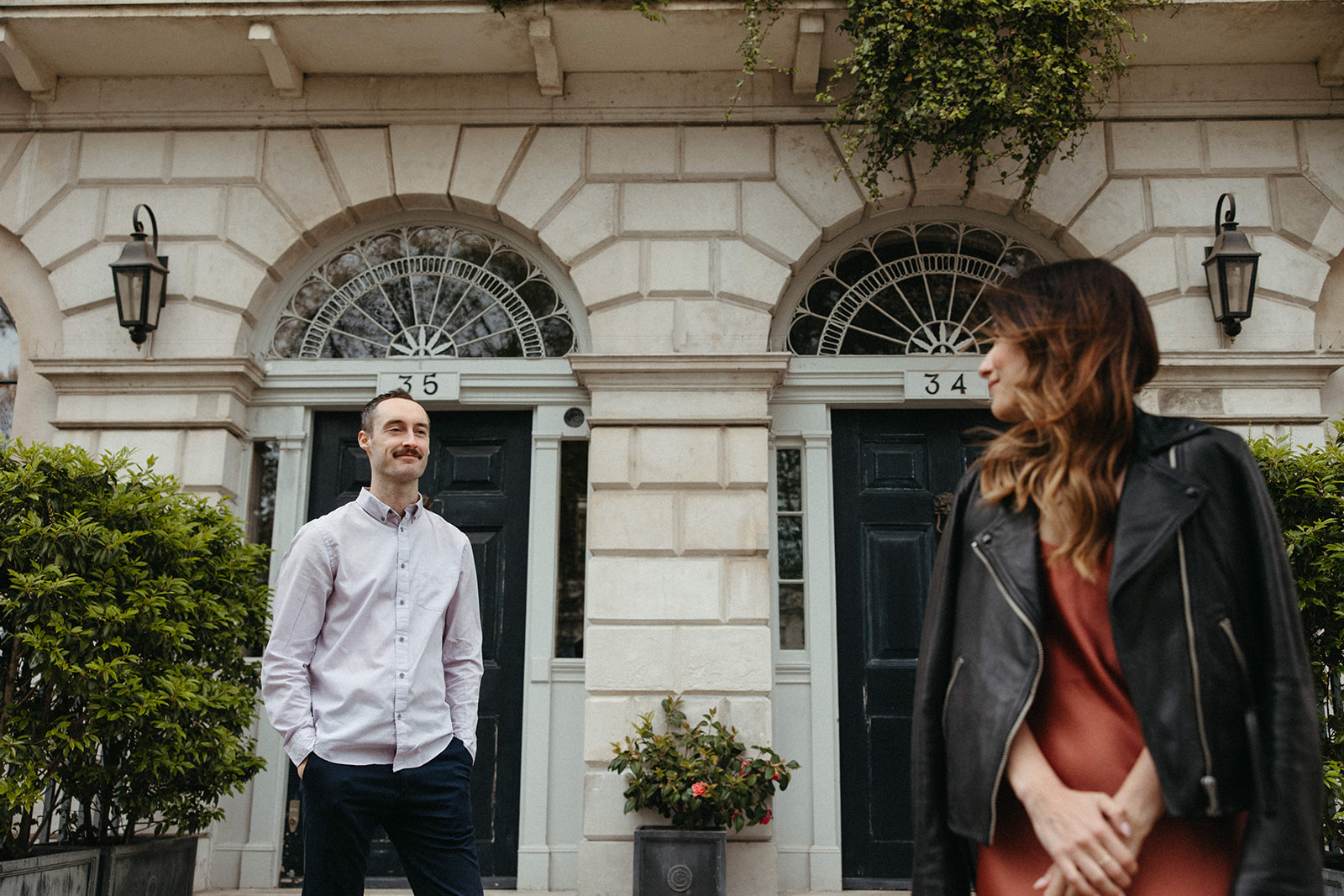 A man dressed in a long-sleeve polo shirt and dark chino pants admires his fiancée during a London engagement session. 