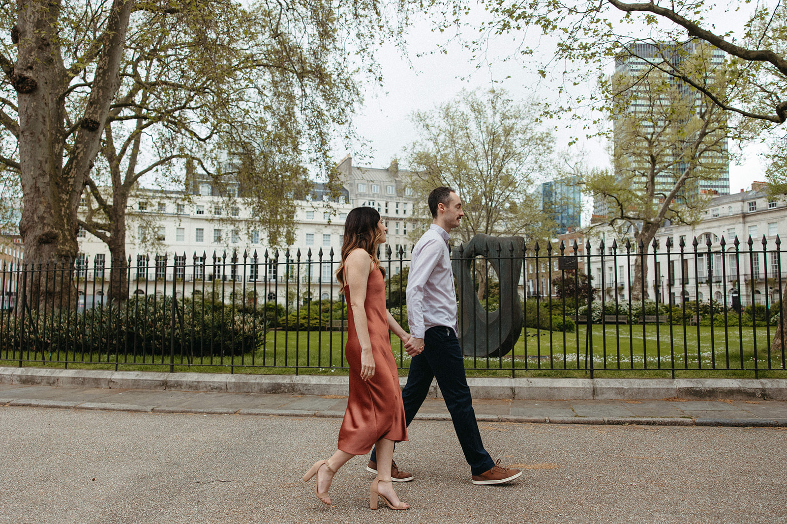 A couple holds hands while walking past a park in London wearing city engagement session outfits. 