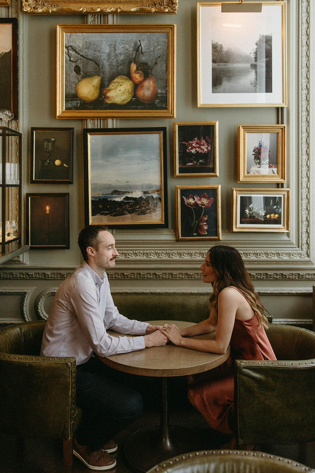 An engaged couple holds hands while sitting in a London restaurant decorated with vintage artwork in gold frames. 