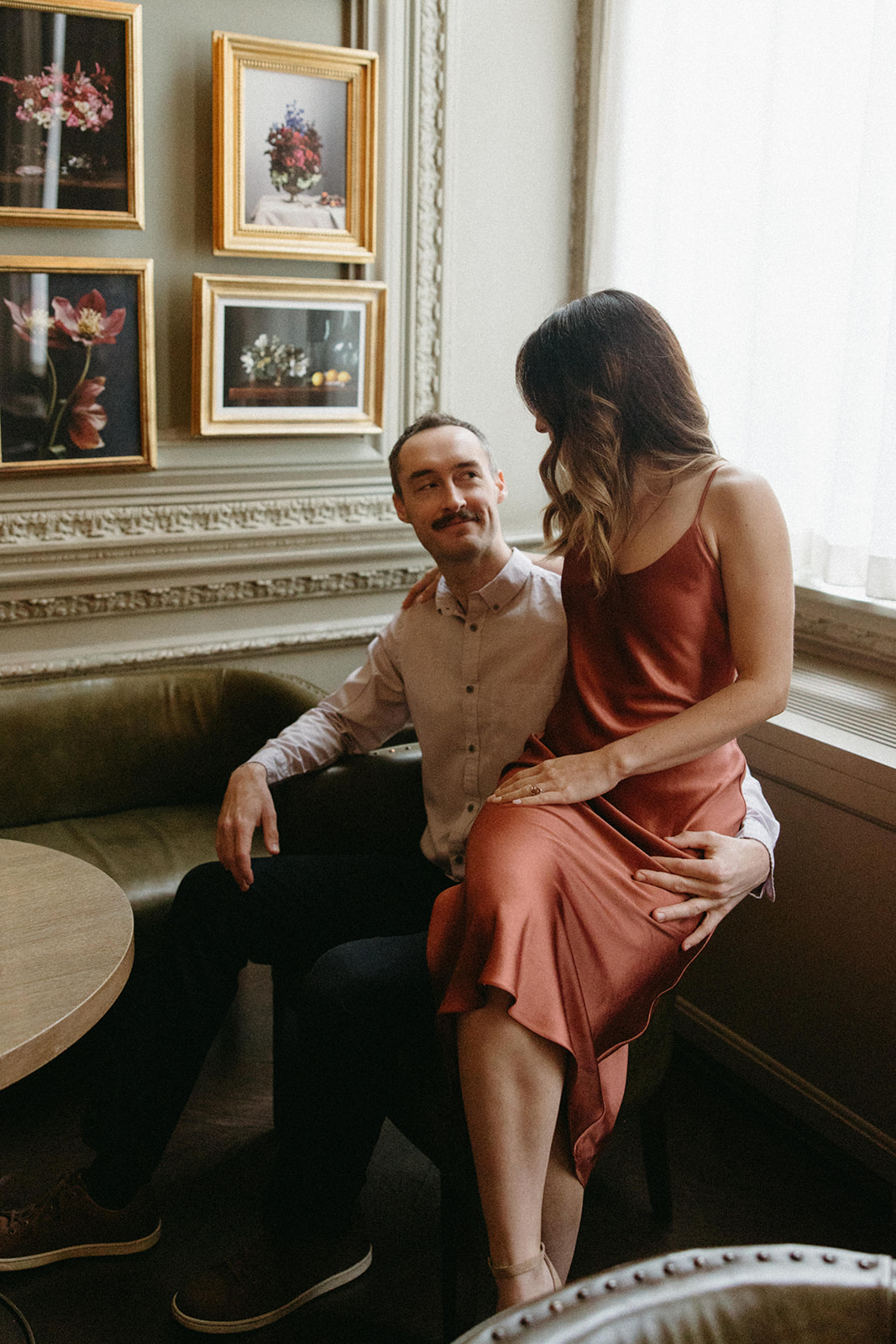 A woman sits on her fiancé's lap in an upmarket London restaurant during their luxury engagement session. 