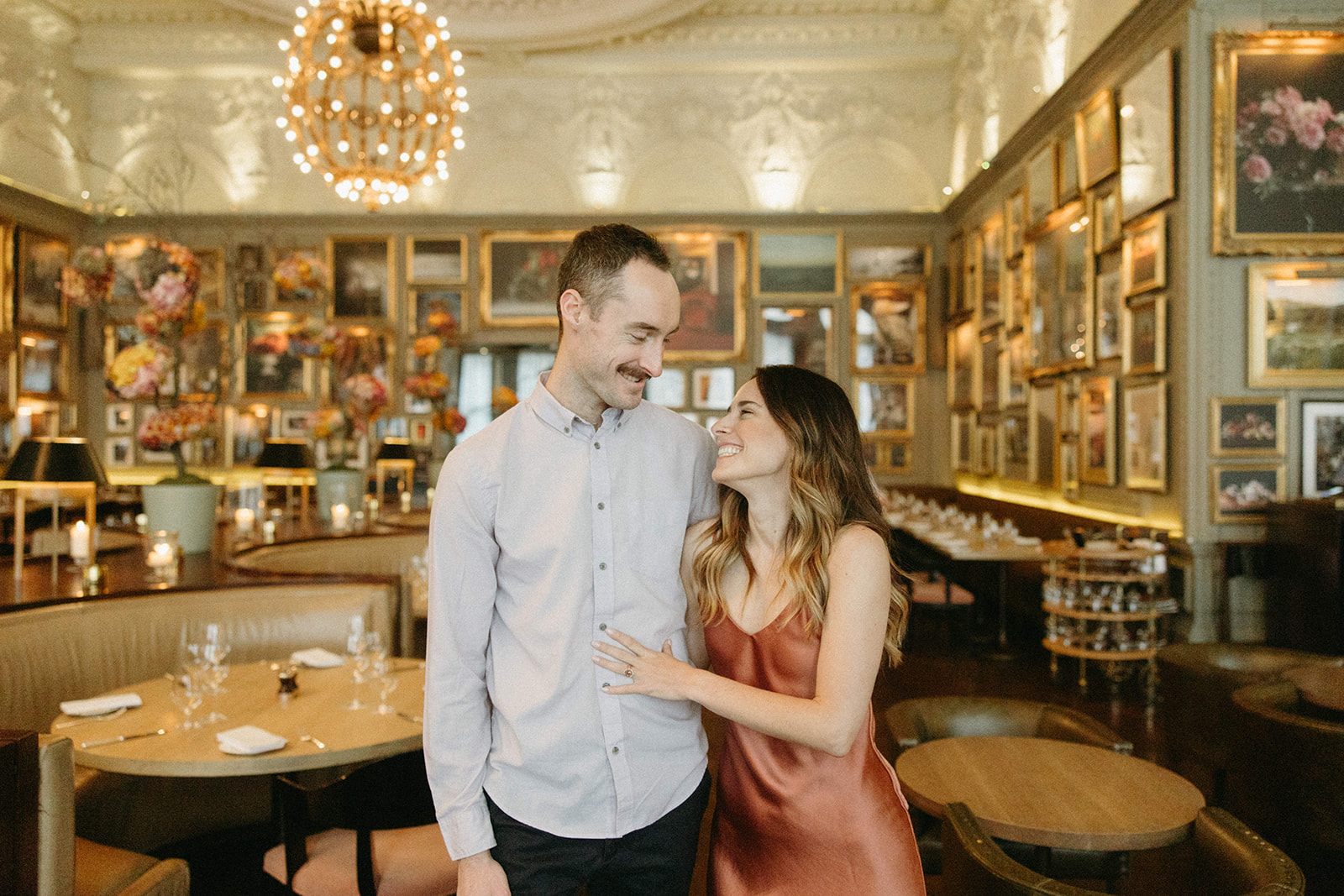 An engaged couple dressed in elevated engagement outfits embrace while laughing in a romantic London restaurant.