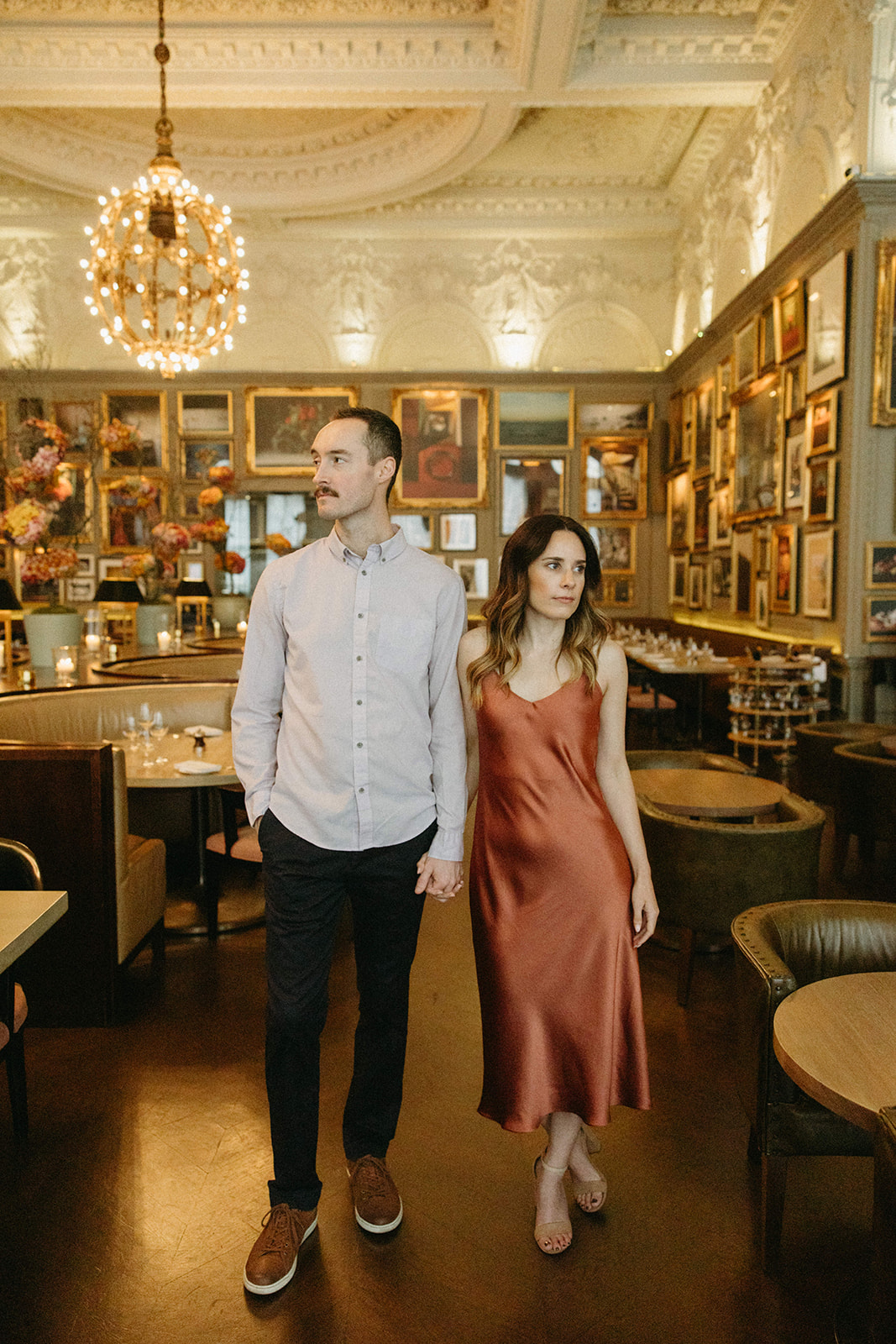 A couple looks opposite ways while holding hands in an elevated tavern decorated with vintage artwork and chandeliers. 