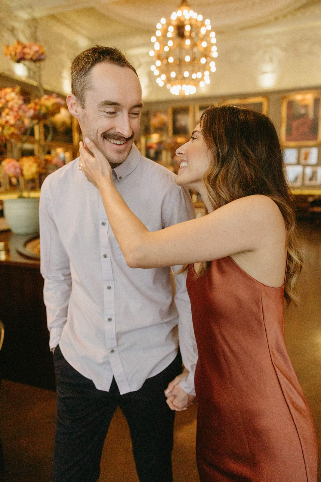 A woman holds her fiancé's face in a romantic London restaurant for their old money aesthetic engagement session. 