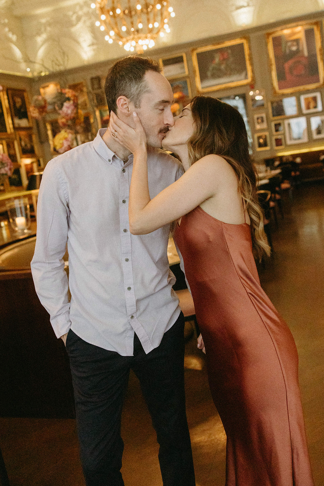 A couple kisses in a charming historical restaurant decorated with chandeliers and vintage artwork in gold frames. 