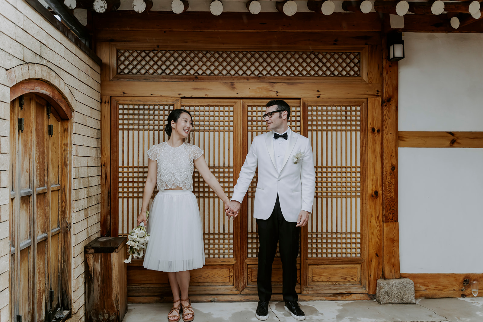 A hanok wedding ceremony with a bride and groom holding hands in front of a traditional wooden door.