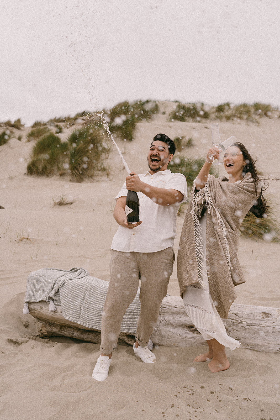 A couple wearing white at their engagement session in Cannon Beach Oregon shot on film and popping champagne