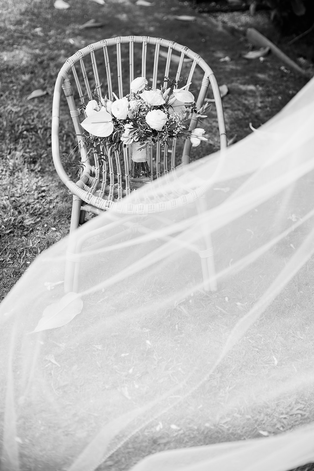 A bridal bouquet rests on a woven chair with a flowing white veil draping onto the grass Wedding at Na ‘Āina Kai