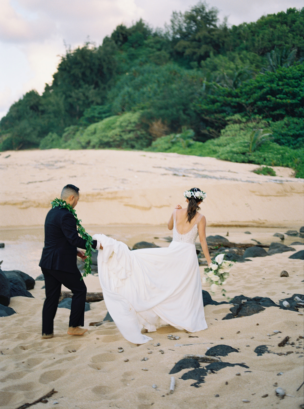 Bride and groom walking on a beach with the groom holding the wedding dress trail at Na Aina Kai