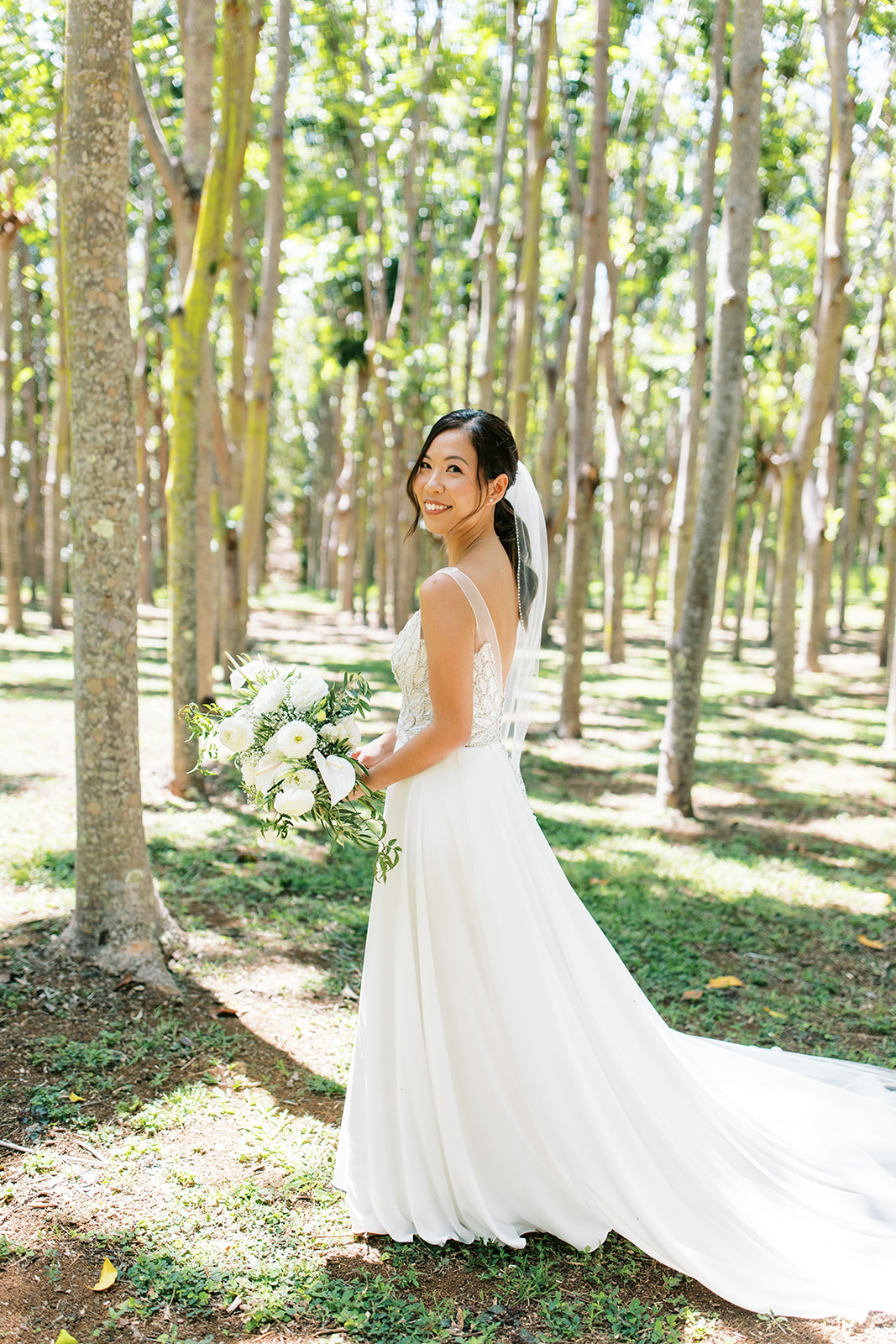 Bride holding a bouquet and smiling in a sunlit forest captured by Oahu Wedding Photographer
 