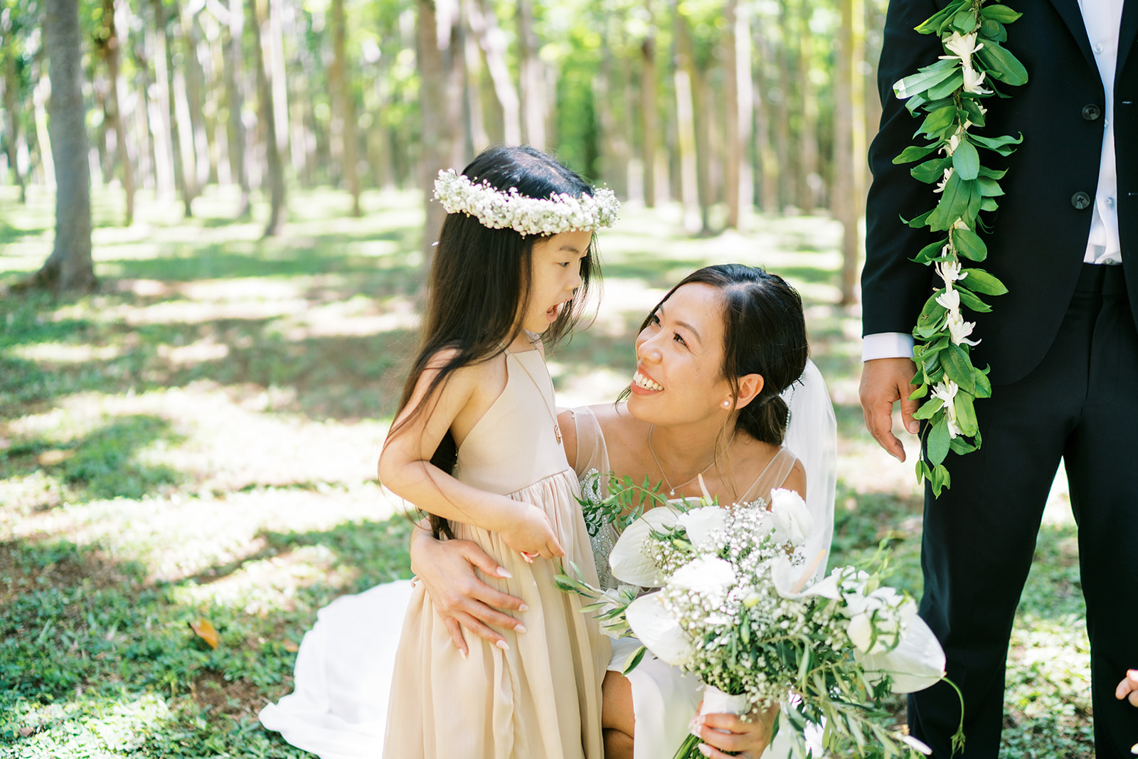 A bride smiles at a young flower girl while holding a bouquet, with a groom partially visible to the right.