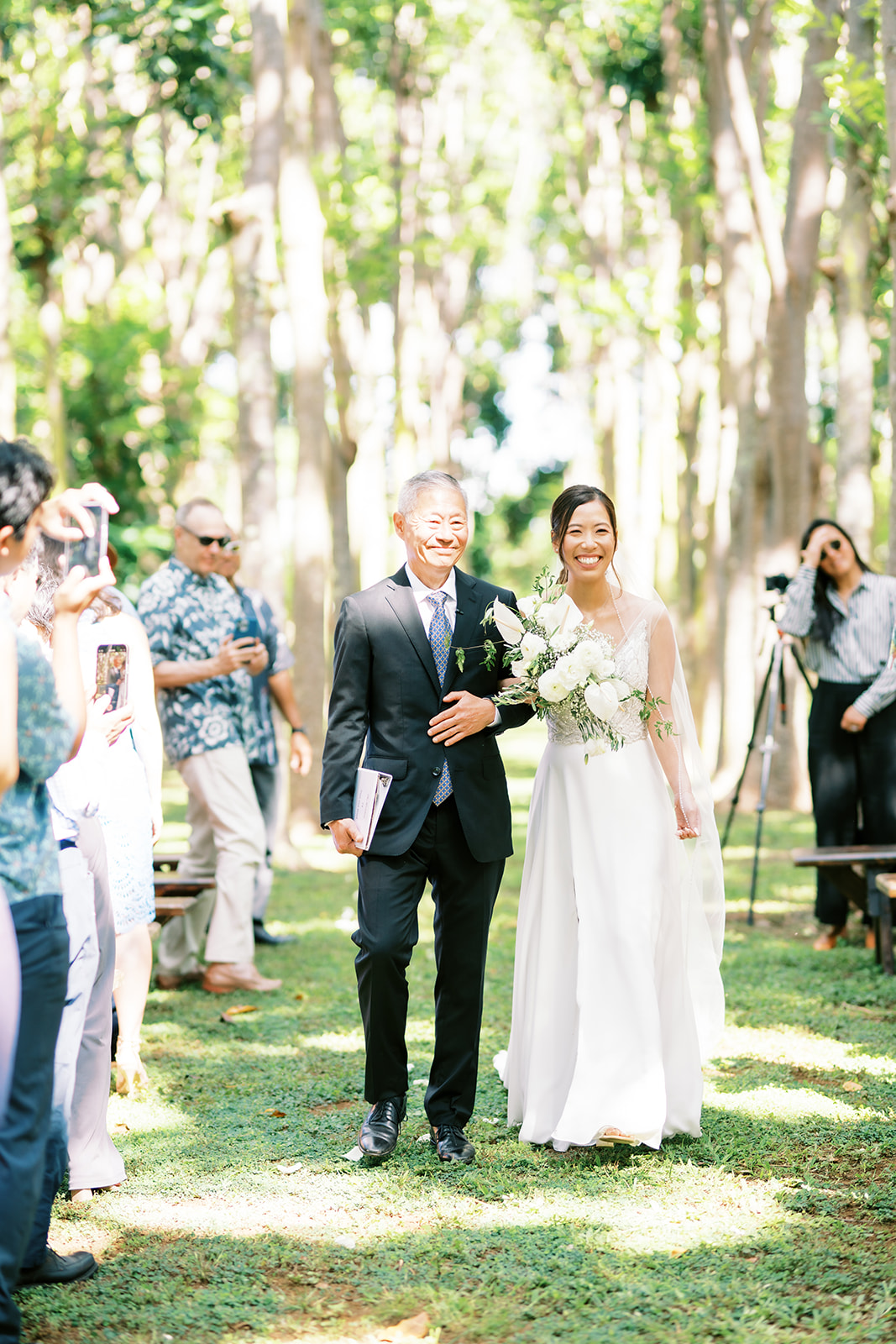 A bride walking down the aisle with her father, both smiling, as guests look on captured by Oahu Wedding Photographer