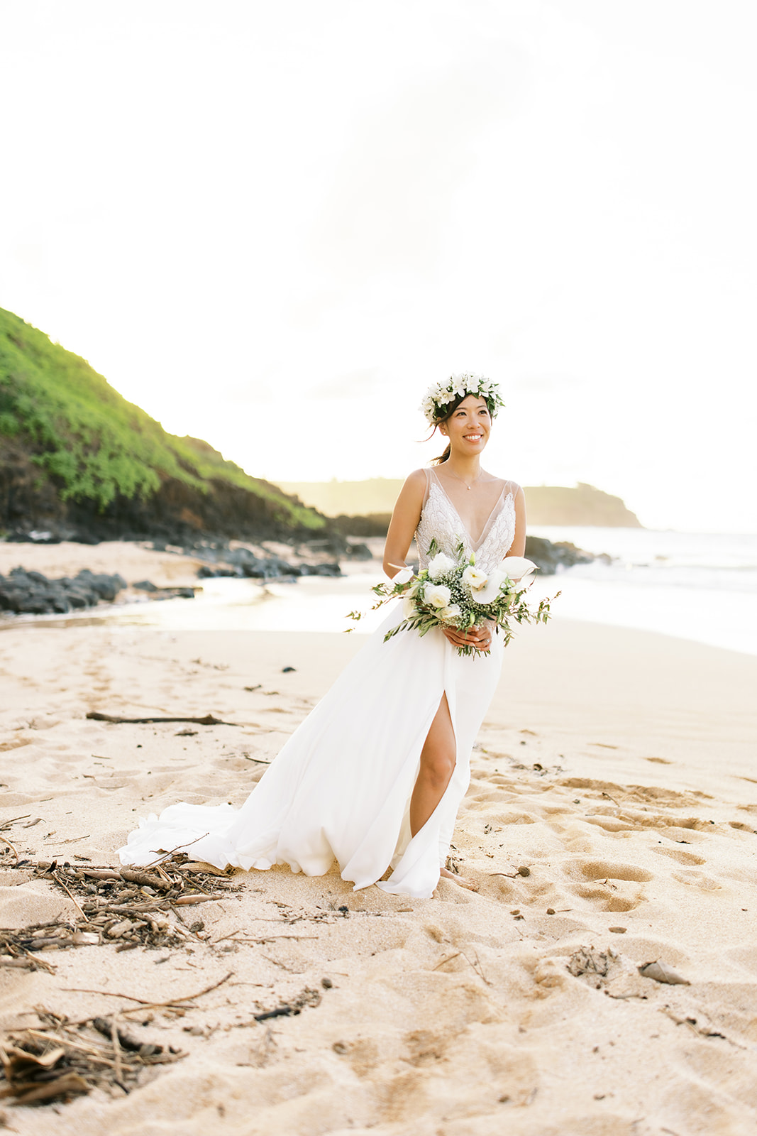 Bride in white dress and floral wreath standing on a beach at Na ‘Āina Kai Botanical Gardens captured by Megan Moura