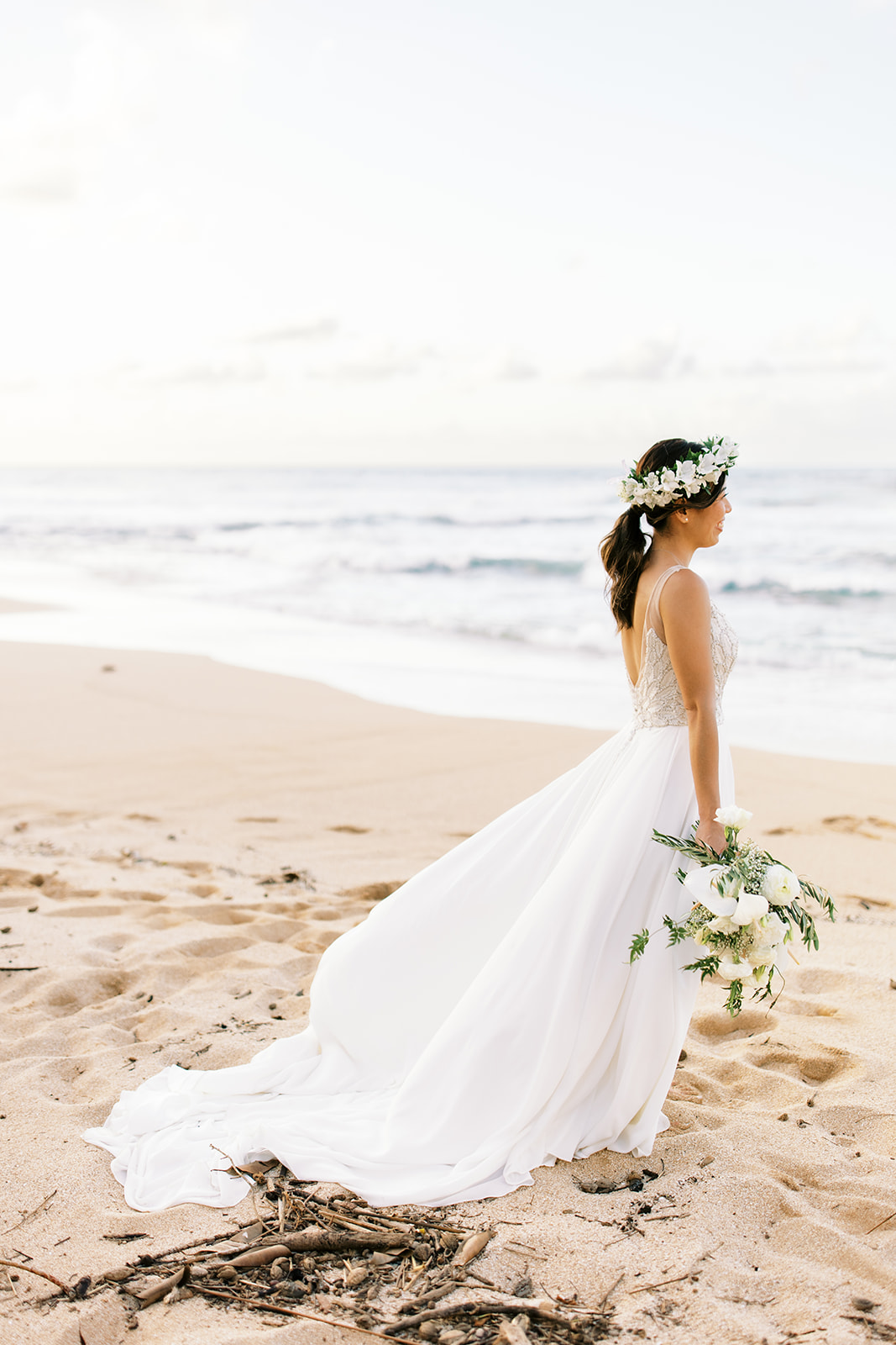 A bride in a white dress holding a bouquet and wearing a floral headpiece walking on the beach at Na Aina Kai