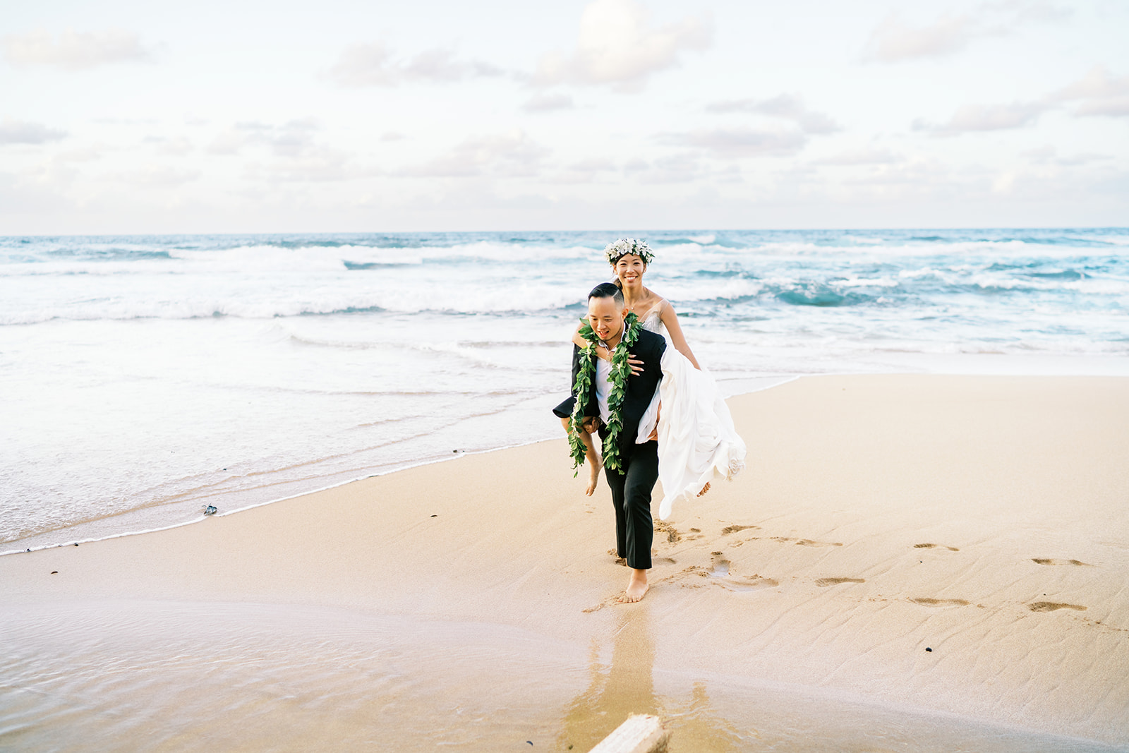 A couple dressed in wedding attire walks barefoot on a sandy beach captured by Oahu Wedding Photographer