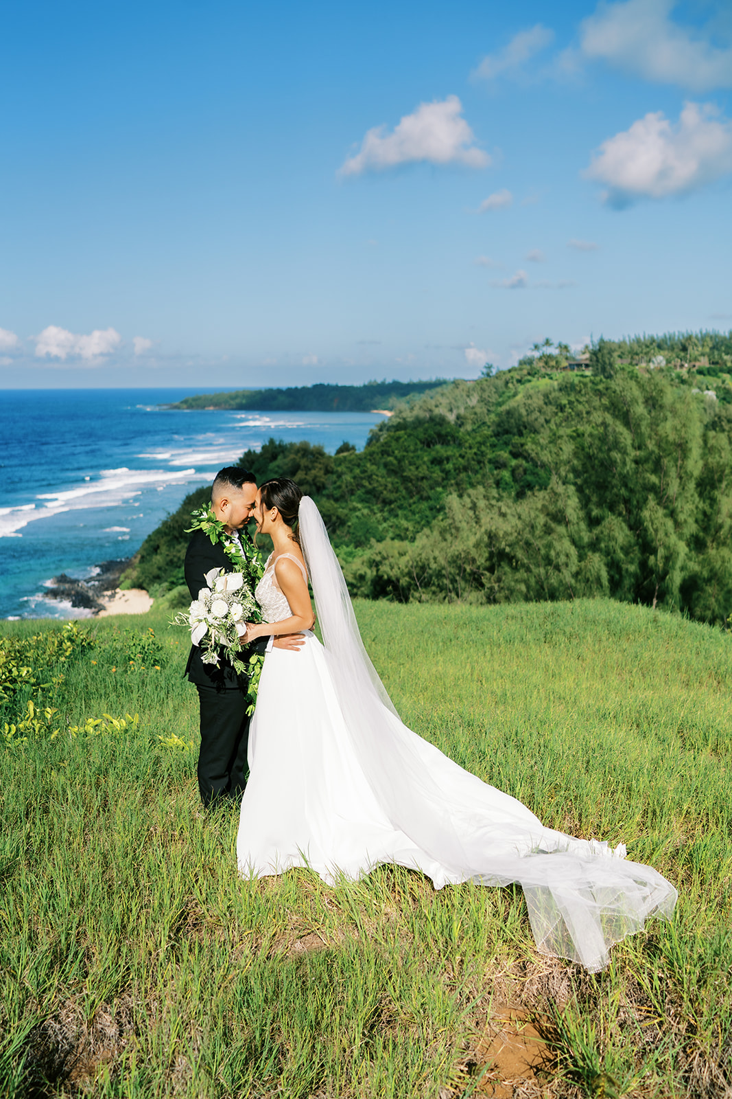 A couple looking at each other, standing in a grassy field with a scenic coastline captured by Oahu Wedding Photographer