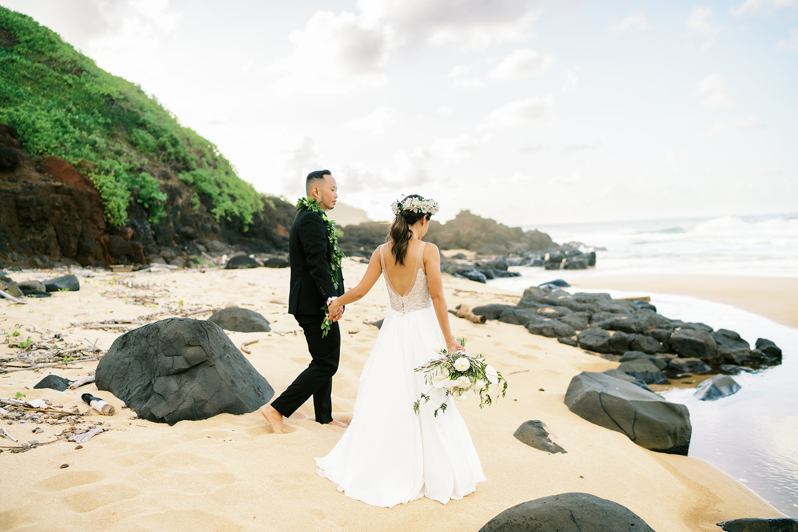 A couple in wedding attire holding hands on Oahu beach with rocks captured by Oahu Wedding Photographer