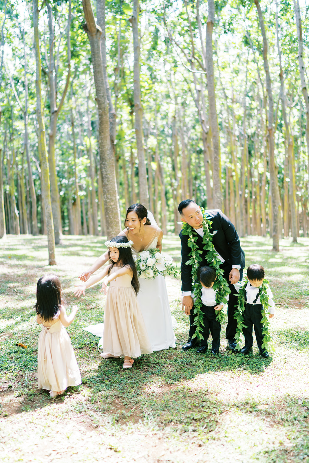 A family with two adults and three children dressed in formal attire, engaging playfully in a sunlit forest clearing