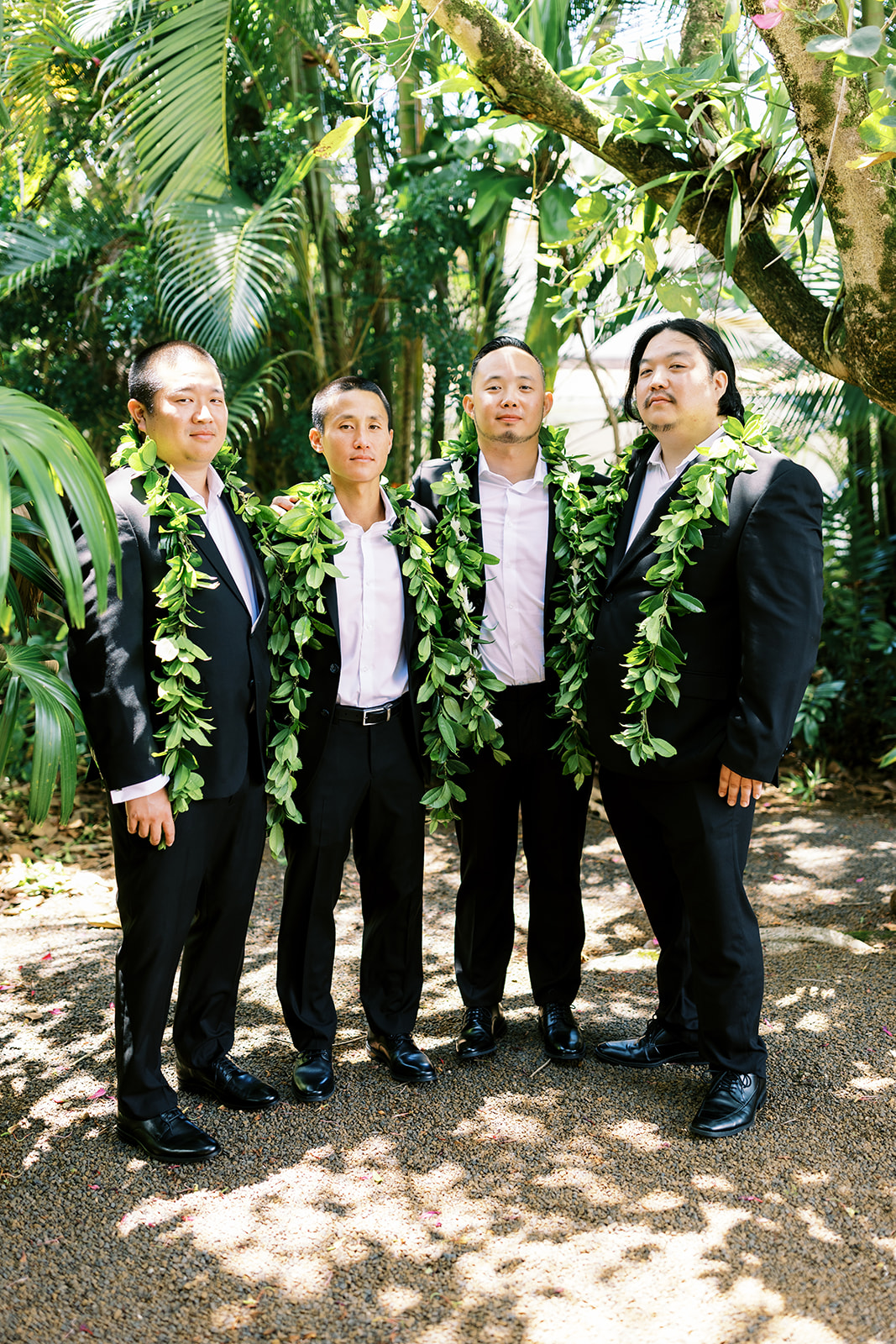 Four men in formal attire with green leaf leis standing outdoors captured by Oahu Wedding Photographer