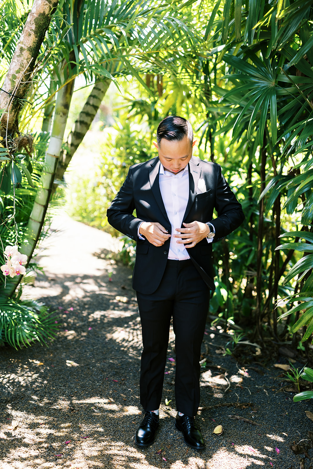 A groom in a black suit fastening his jacket amidst lush greenery captured by Oahu Wedding Photographer