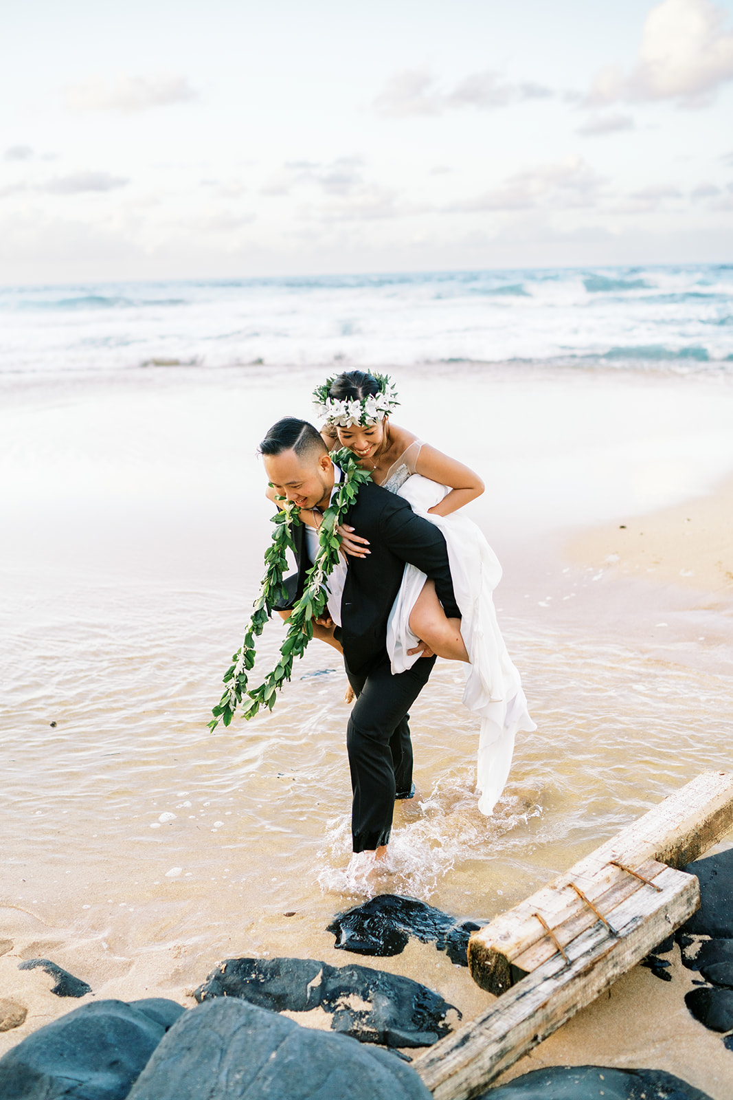 Groom carrying bride on his back along a beach shoreline Wedding at Na Aina Kai captured by Oahu Wedding Photographer