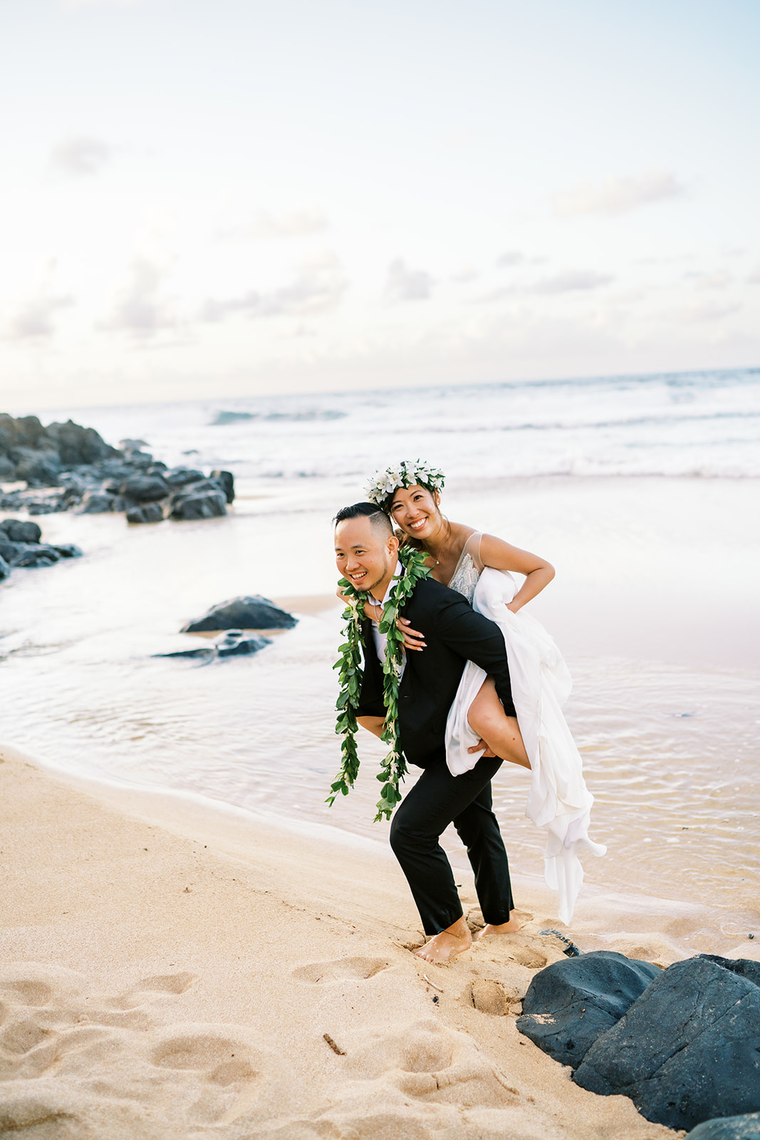 A joyful couple in formal attire, with the man giving the woman a piggyback ride along the shore of Oahu at Na Aina Kai