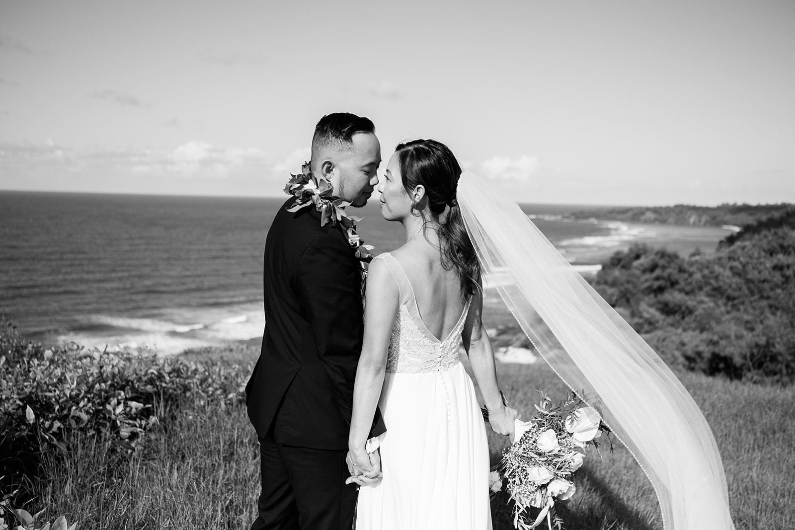 A monochrome photo of a bride and groom share a kiss on a scenic coastline, with the bride's veil caught in the breeze.