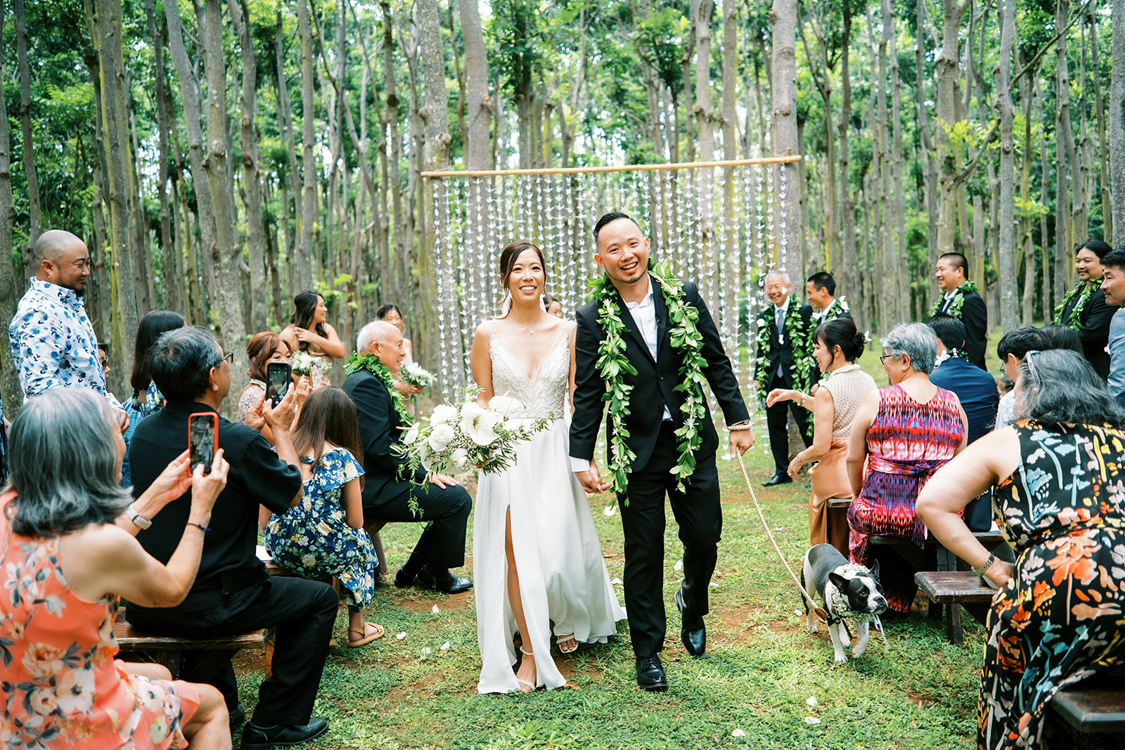 Newlyweds holding hands and smiling as they walk past seated guests in Na Aina Kai captured by Oahu Wedding Photographer