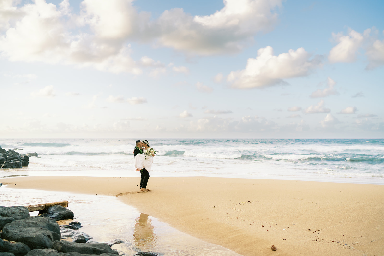 Newlyweds on Oahu beach holding a bouquet of flowers, with waves in the background captured by Oahu Wedding Photographer