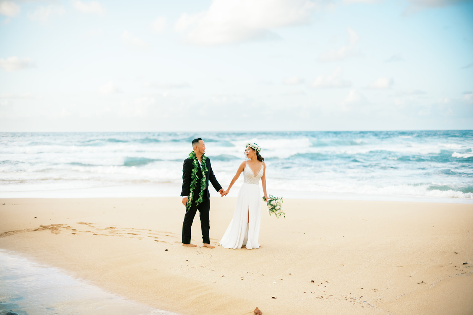 Newlyweds in wedding attire strolling on a sunny beach Wedding at Na Aina Kai captured by Oahu Wedding Photographer
