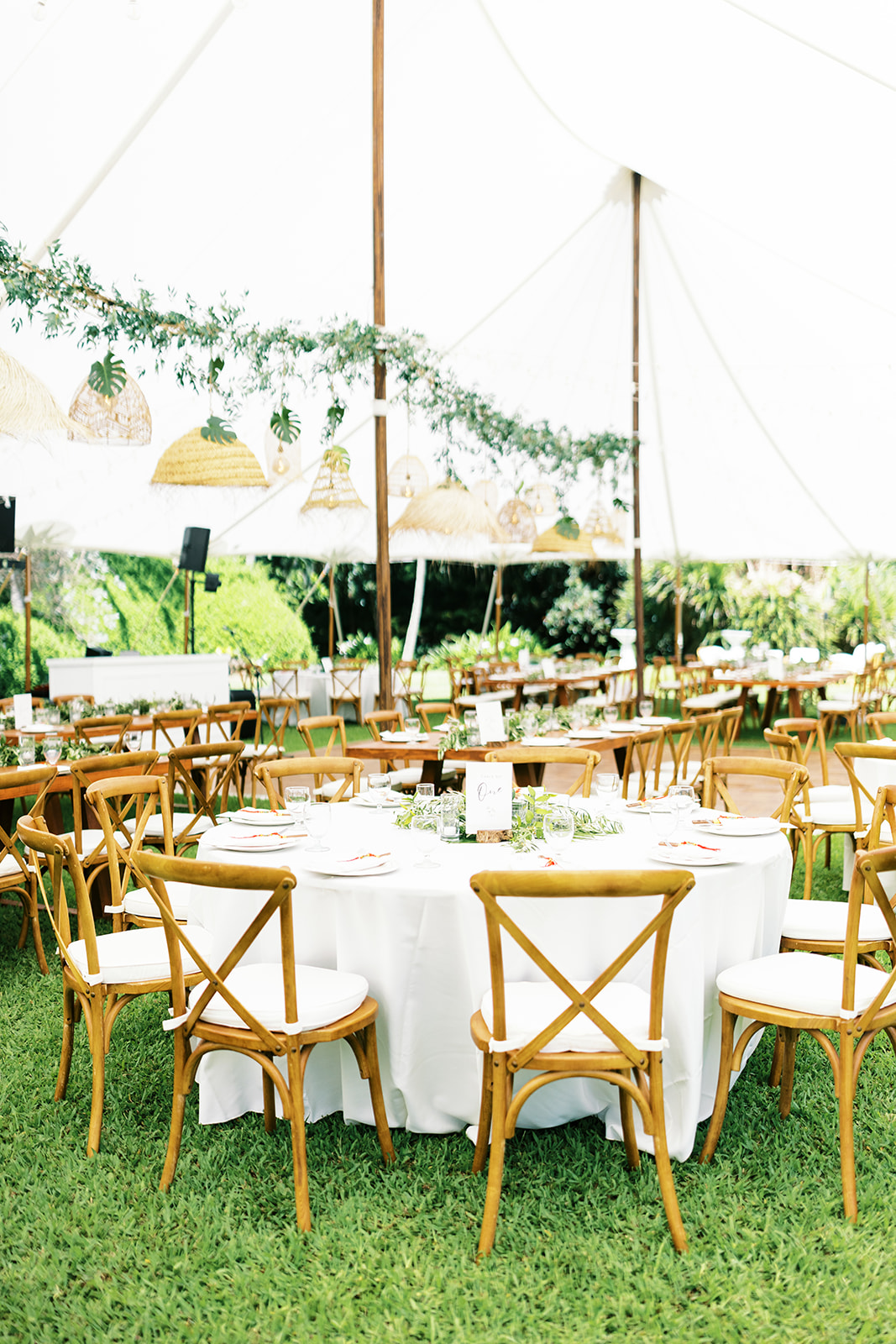 Outdoor wedding reception setup with bamboo chairs, white tablecloths, and hanging greenery under a white tent.