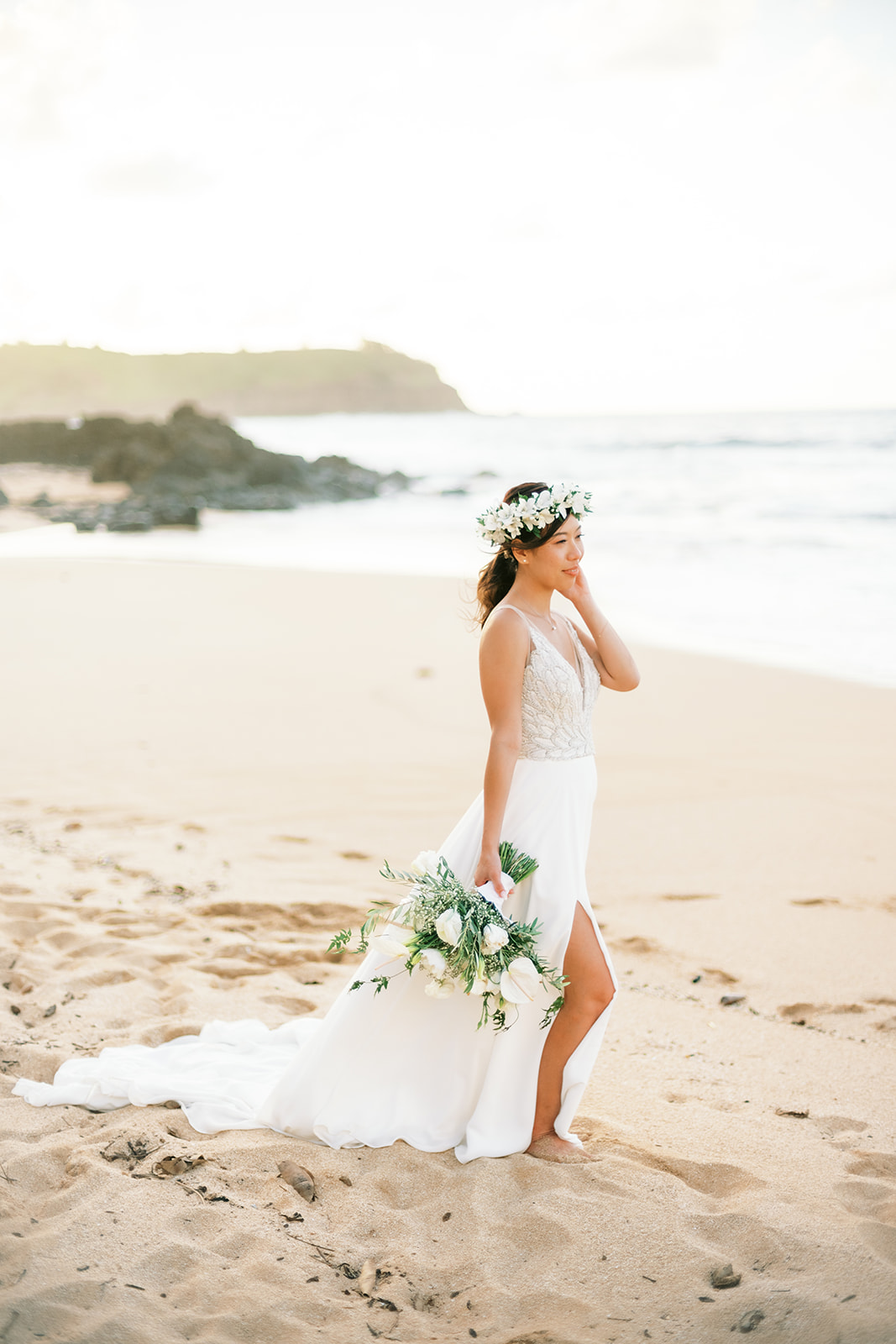portrait of a bride in a white dress holding a bouquet on a sandy beach fixing her air, looking to the side with a smile