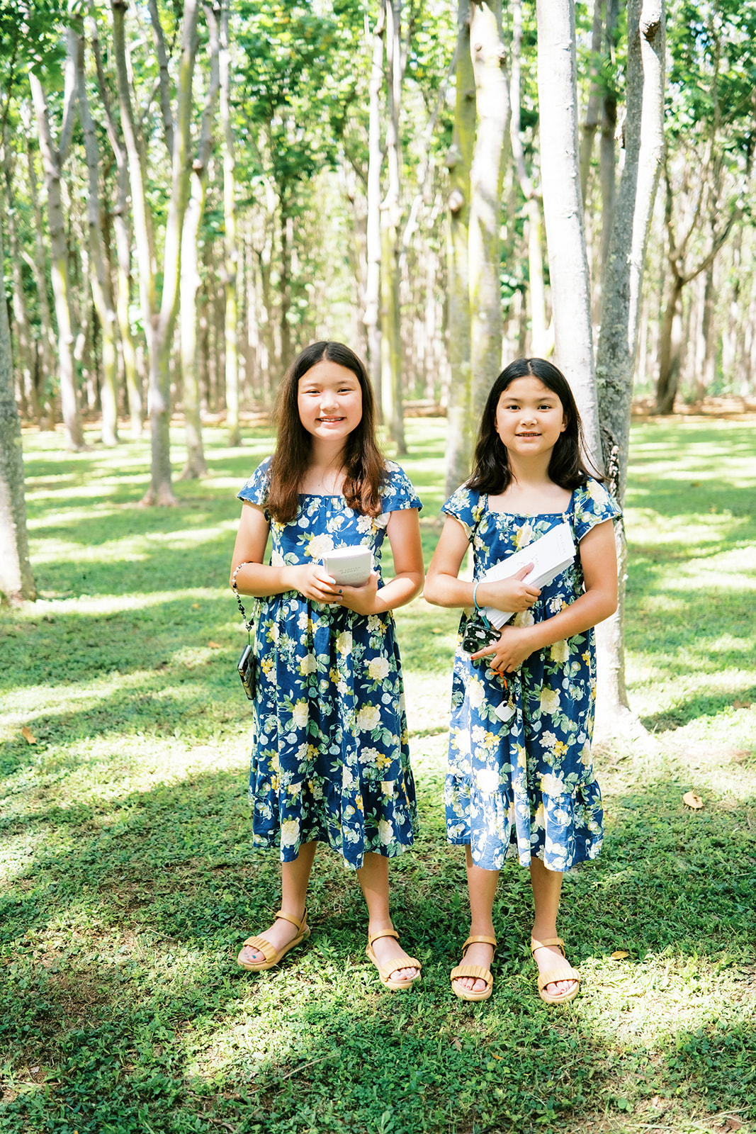 Two girls in matching floral dresses standing in a forest clearing Wedding at Na ‘Āina Kai captured by Megan Moura