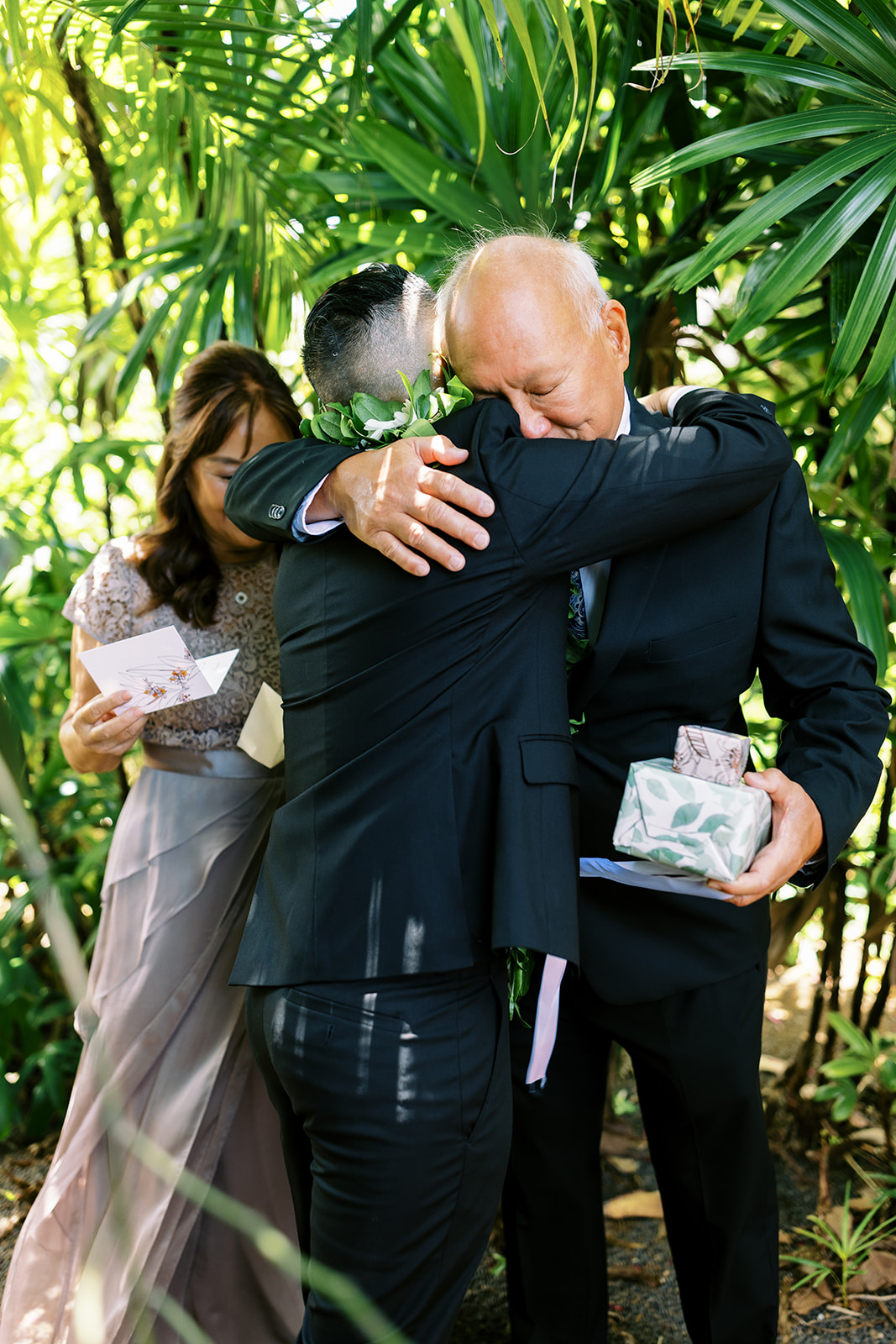 Two men embracing at a wedding celebration, with a woman holding a card and a gift in the background Wedding at Na Aina 