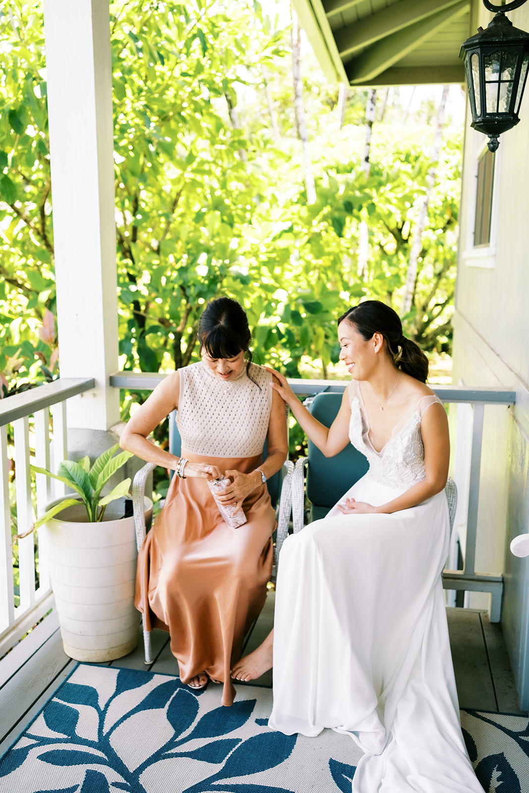 Two women sharing a joyful moment on a porch, one in a bridal gown and the other in a formal dress Wedding at Na ‘Āina K