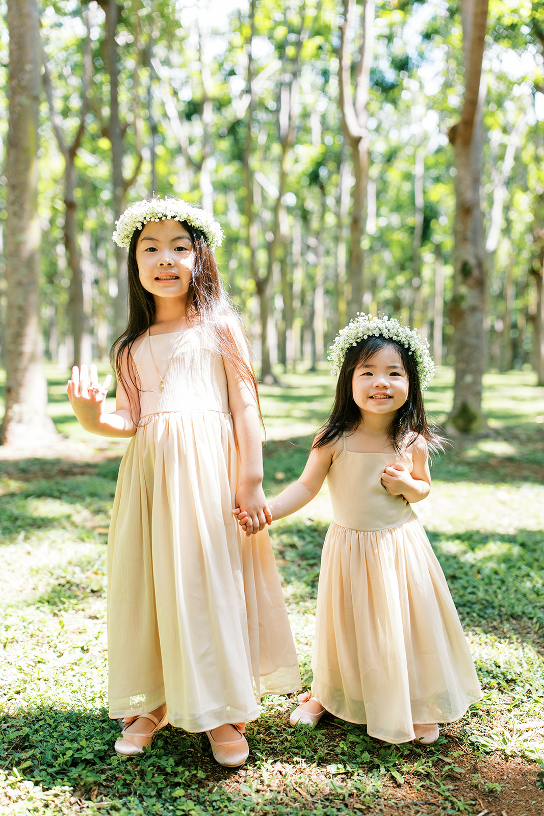 Two young girls in matching dresses and floral crowns standing in a sunlit forest Wedding at Na ‘Āina Kai Botanical