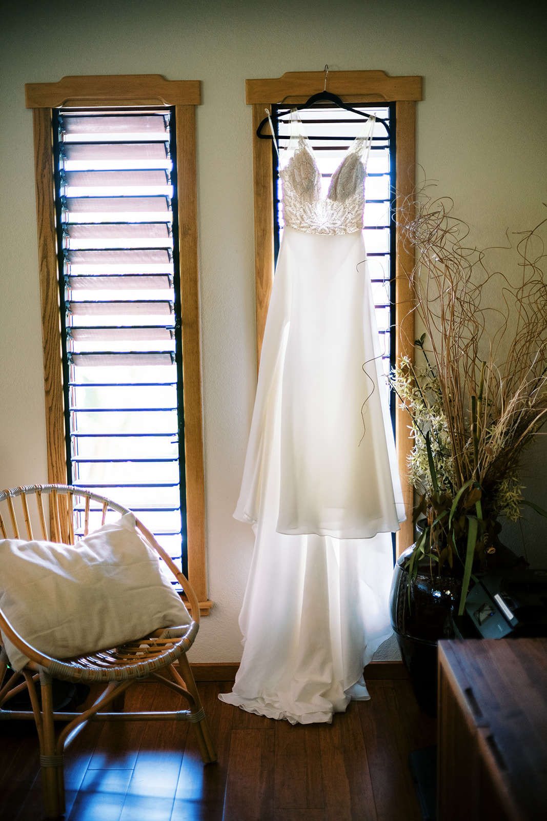 A wedding dress hanging by a window, with a chair and vase nearby Wedding at Na ‘Āina Kai Botanical Gardens