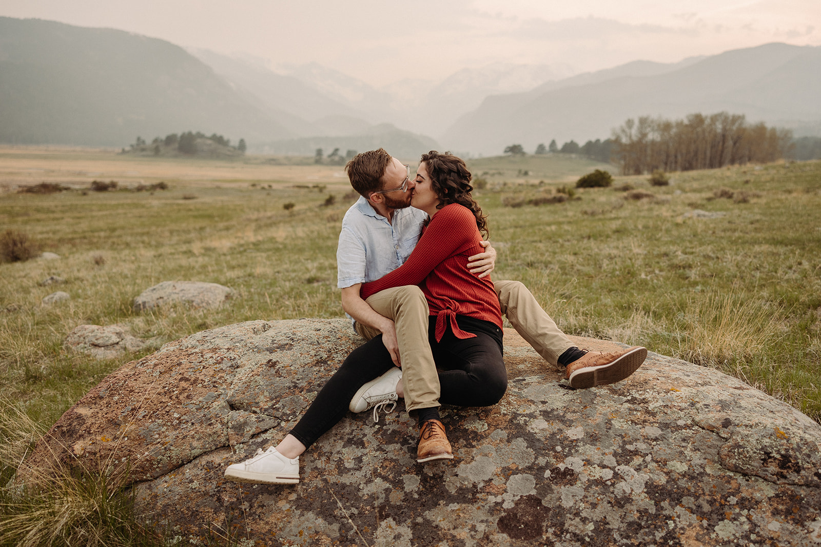 A man and woman sitting on a big rock, kissing with mountains in the background