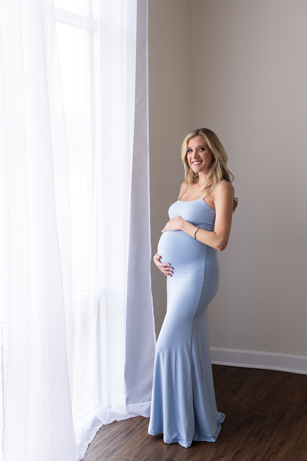 Mom to be stands next to a big window wearing a baby blue maternity dress