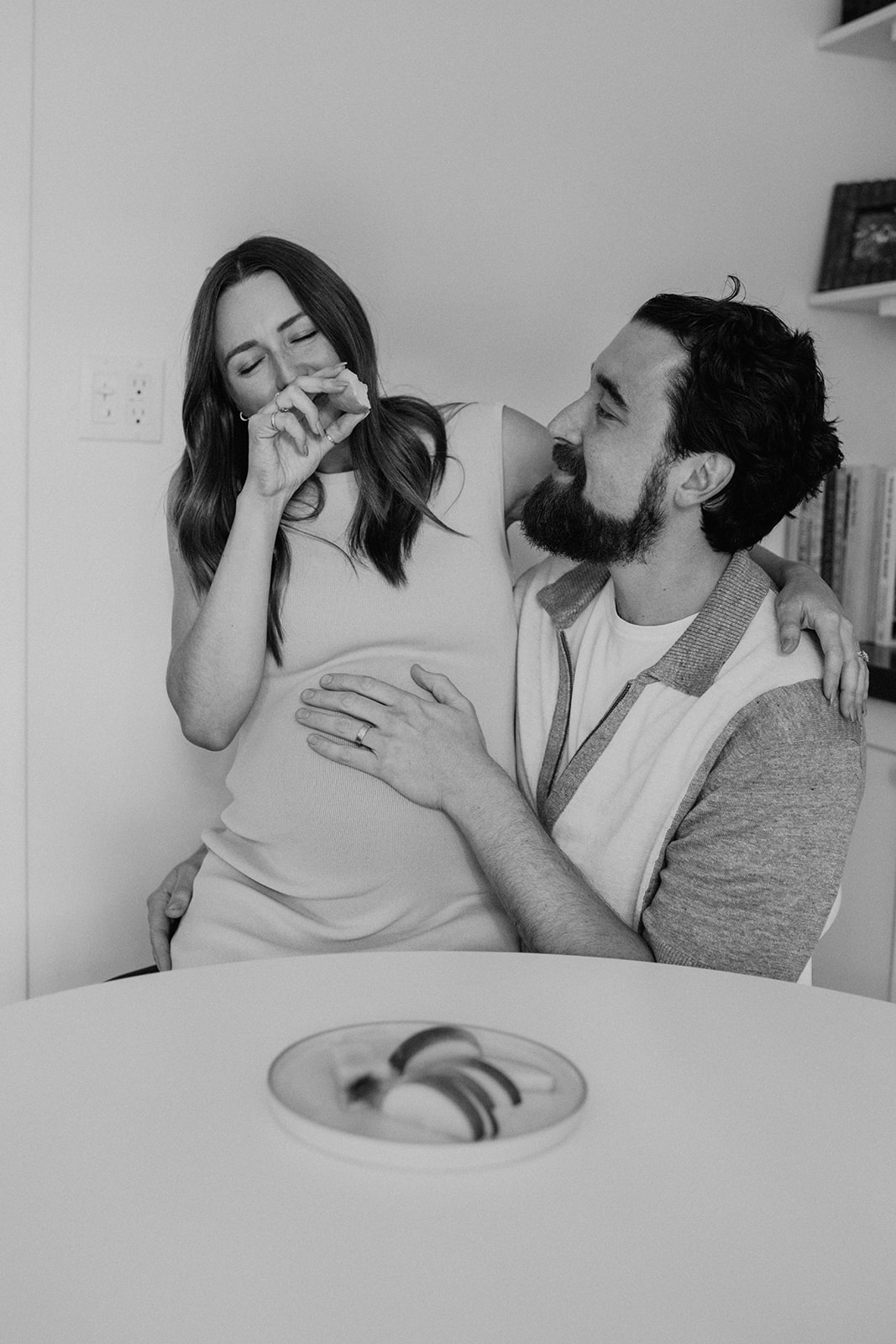 black and white photo of a pregnant woman sitting on her partner's lap eating an apple