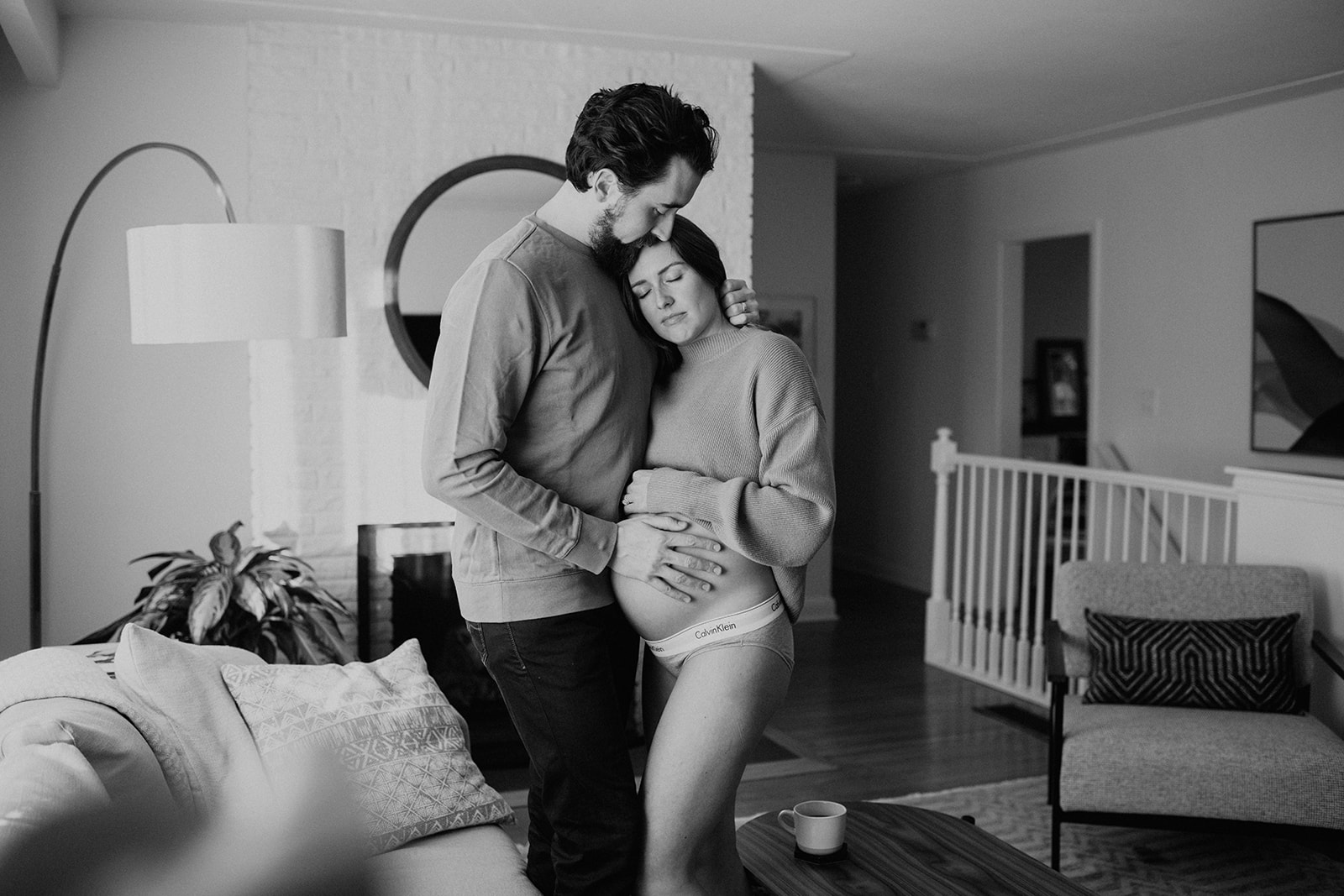Husband and wife hug close in their living room for a black and white maternity photo