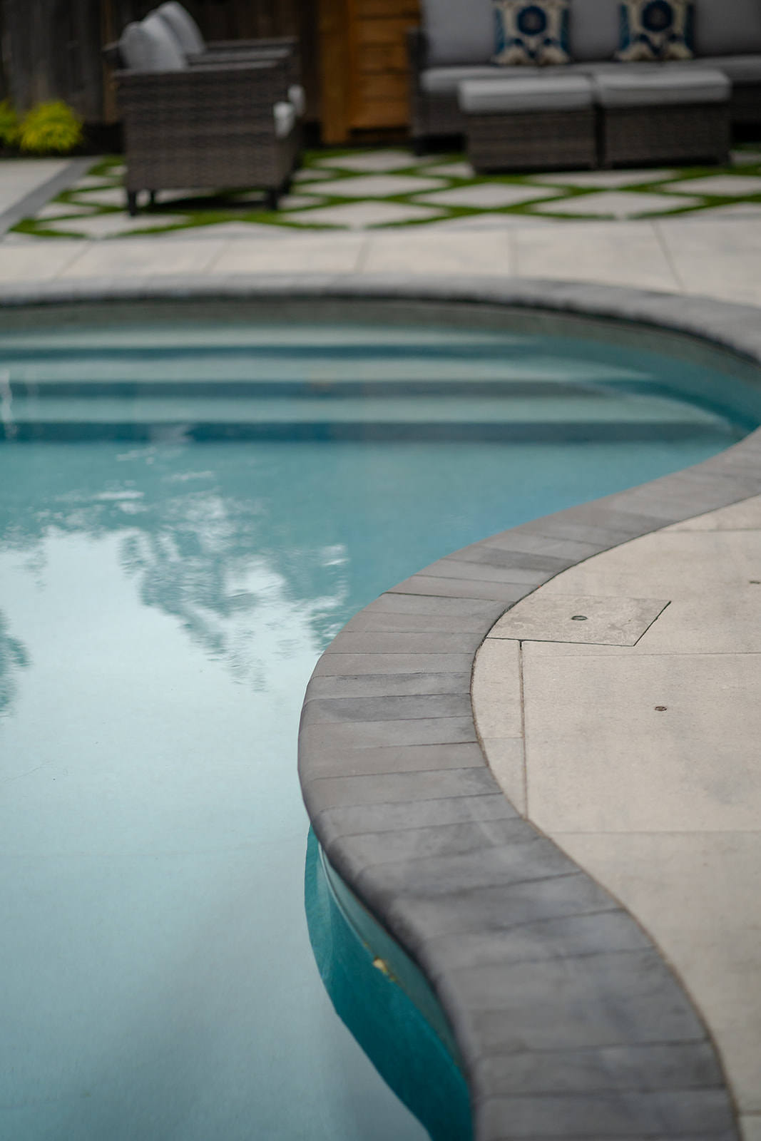 Interlocking patio stones lining the inground pool while stairs on the opposite end are leading into the pool.