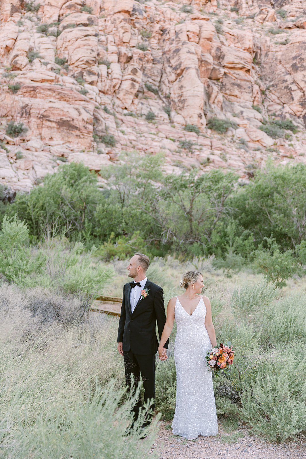 bride and groom hold hands on Red Rock Boardwalk with Red Rock Canyon backdrop. Bride wearing glittering white dress.