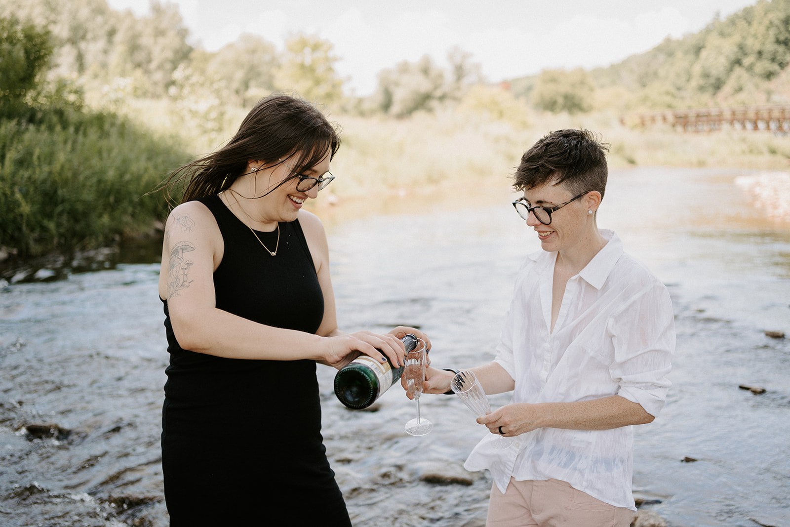 One person pouring champagne into their partners glass while standing in the creek.